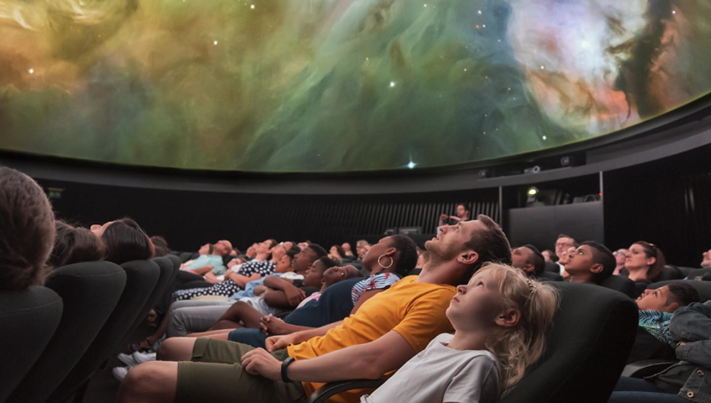 A large group of people, watching a planetarium show. The screen is showing a swirl of a galaxy