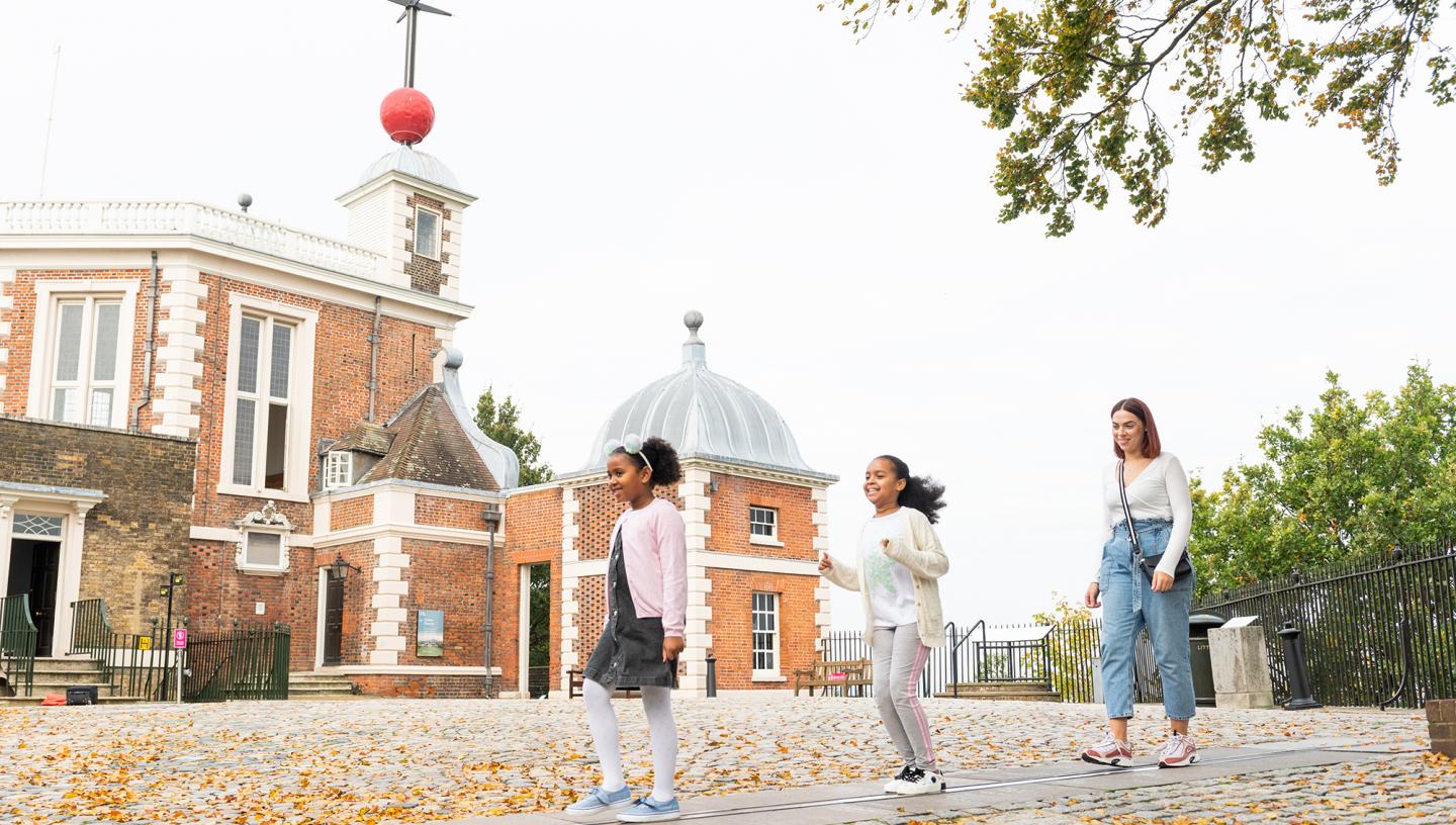 A family play on the Prime Meridian Line in Greenwich, with the historic Royal Observatory building in the background