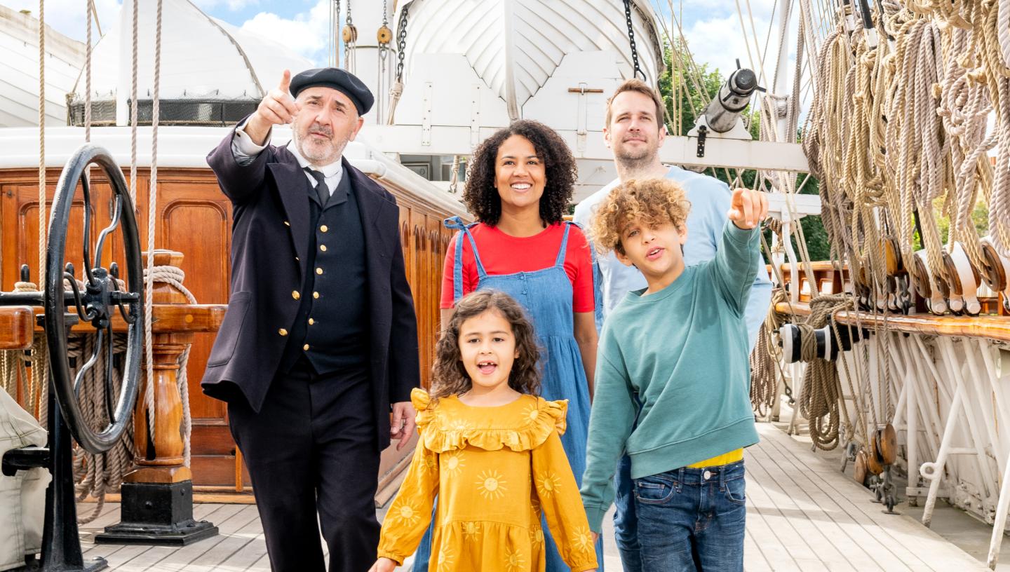 A family walks along the Main Deck of Cutty Sark, accompanied by a character actor dressed as the ship's captain. They are pointing to something behind the photographer, and the ship's rigging and lifeboat frames them in the background