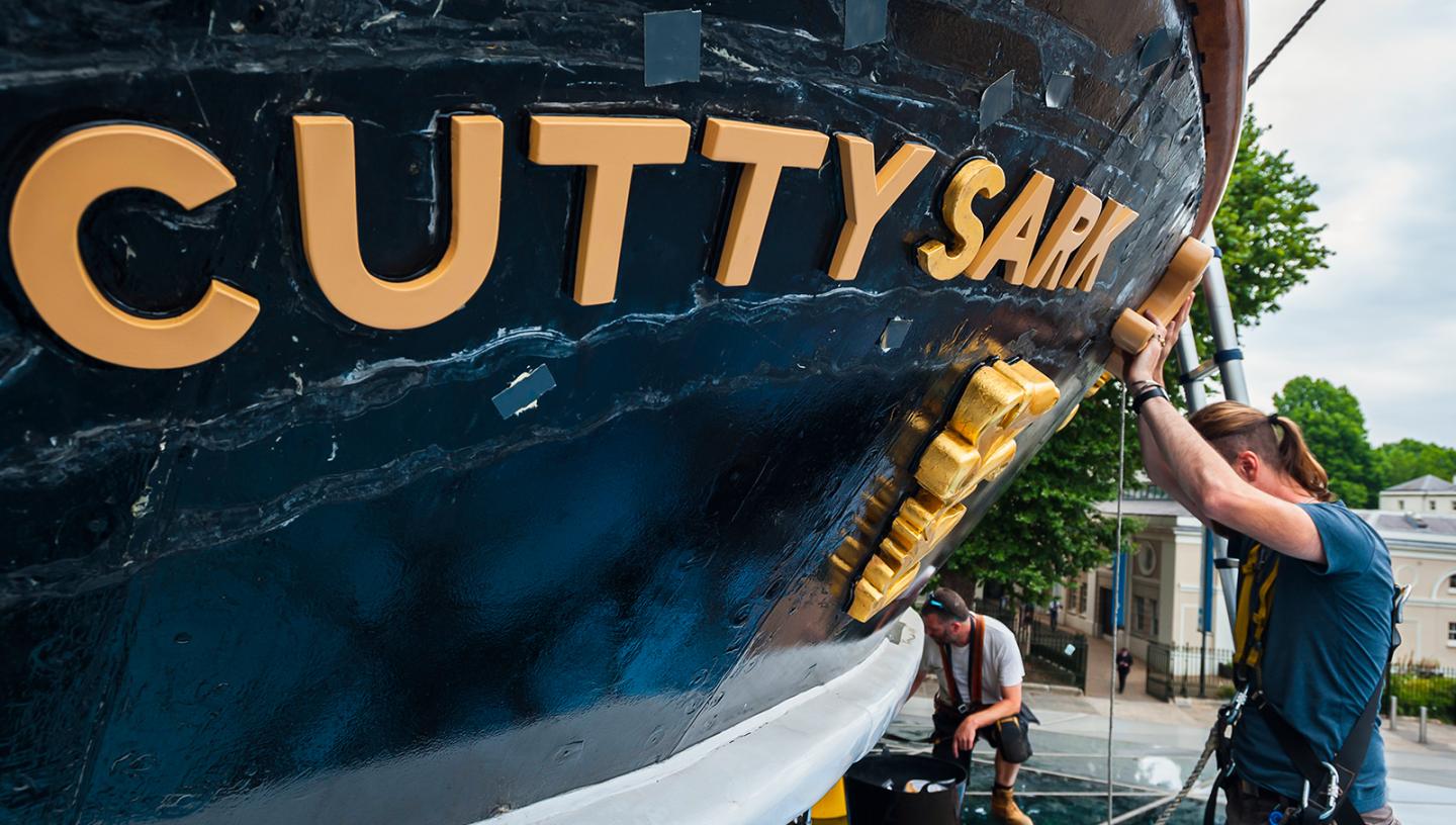 A man in a harness works on historic ship Cutty Sark, with the name of the ship picked out in gold lettering
