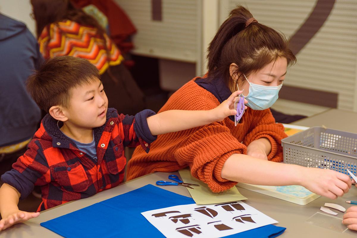 A boy proudly holds up a stencil of a ship while a woman in a face mask directs a craft activity at the National Maritime Museum