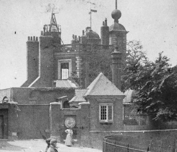 Historic photo of woman standing in front of Shephard Clock in front of observatory wall, with Flamsteed House in the background