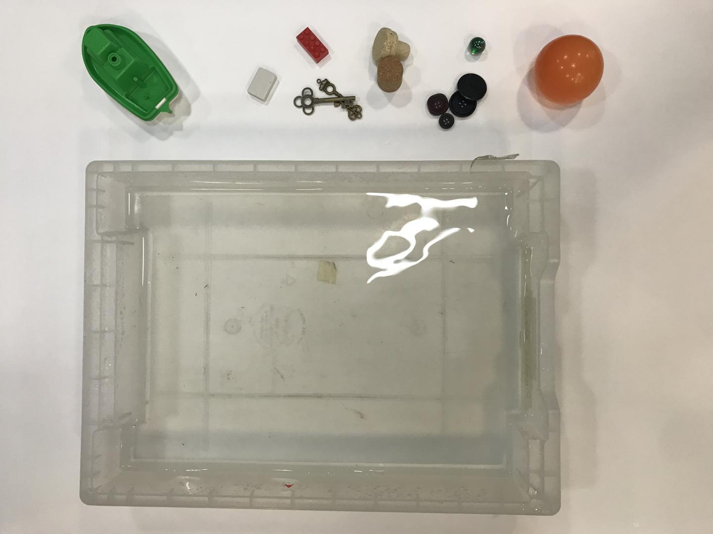 a tray of water with different objects including a toy boat, keys and corks nearby. 