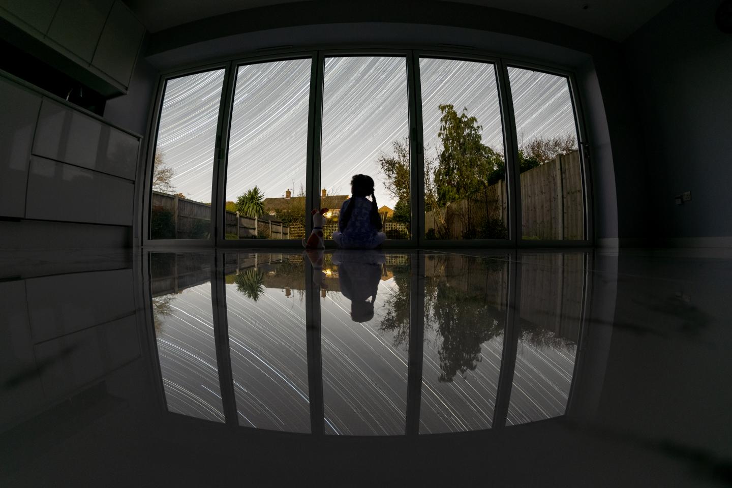 A girl sits behind bi-folding doors overlooking a garden. Star trails cross the sky above