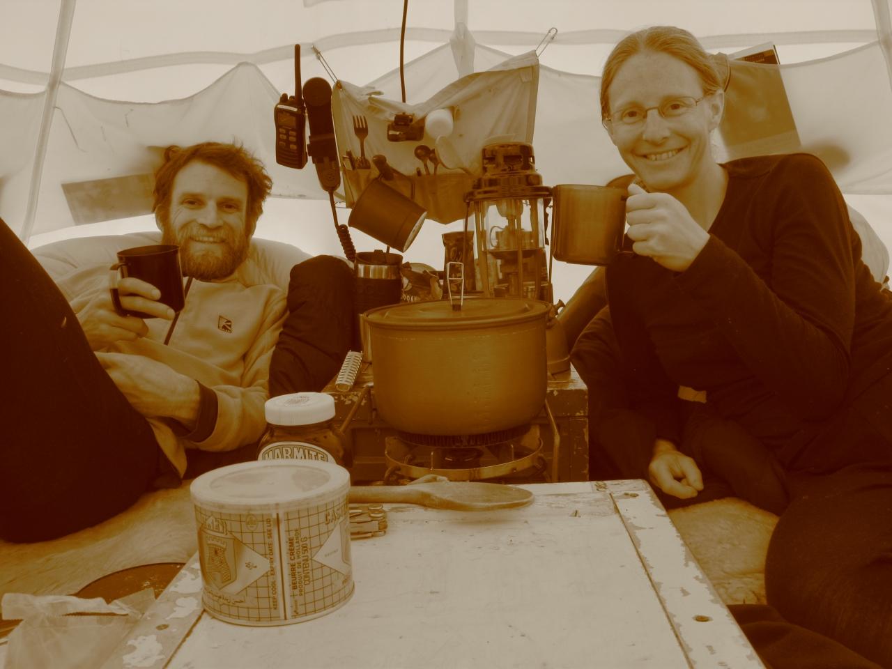 Two people in a tent smile and raise their mugs to the camera. A vintage effect filter gives the photo a brownish tint