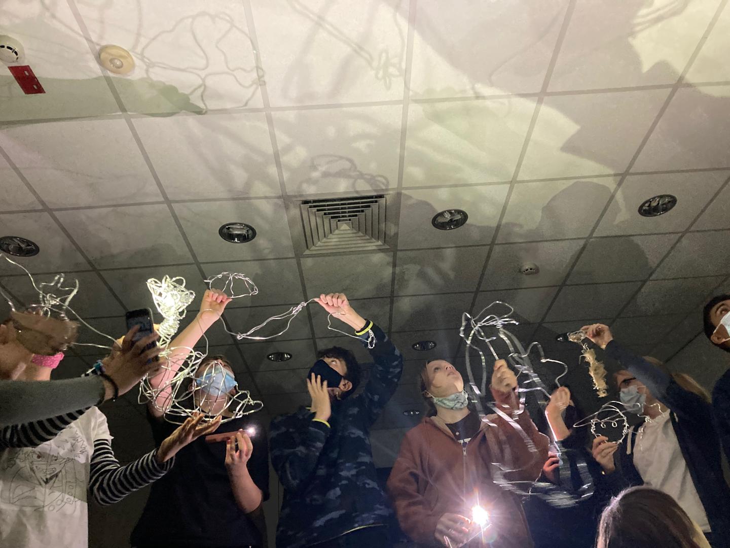 A group of young people looking at shadows on ceiling created by wire sculptures in front of light source