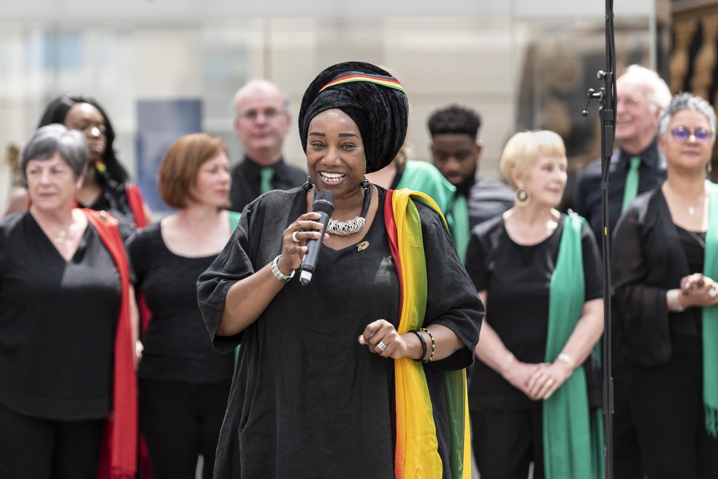 The image shows a choir of people with one woman in front holding a microphone.