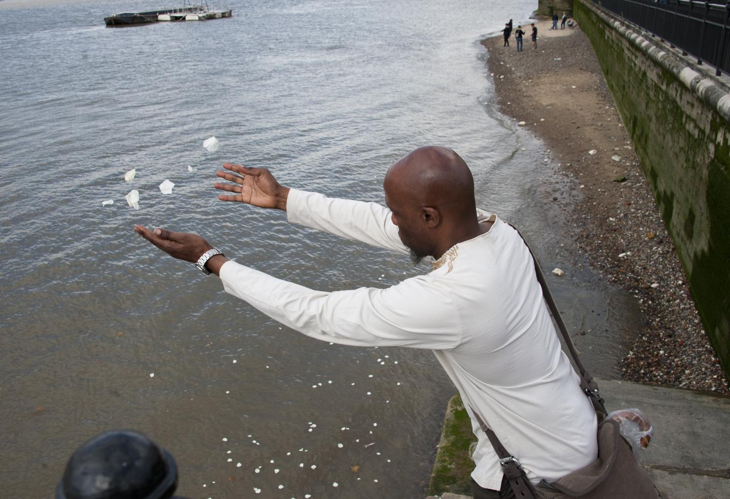 The image shows a man scattering white petals into the River Thames.