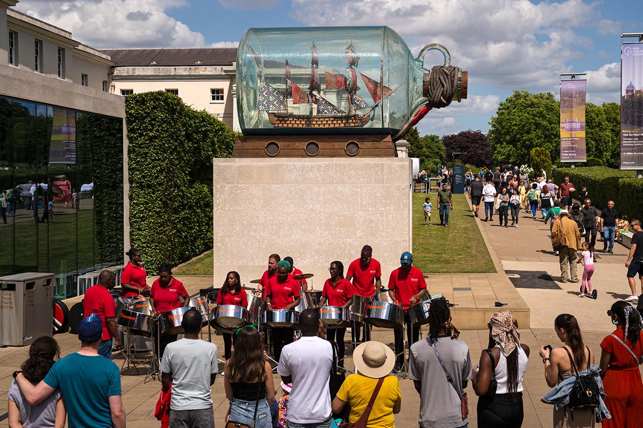 A steel band plays beneath a sculpture of a ship in a bottle outside the National Maritime Museum as a crowd look on