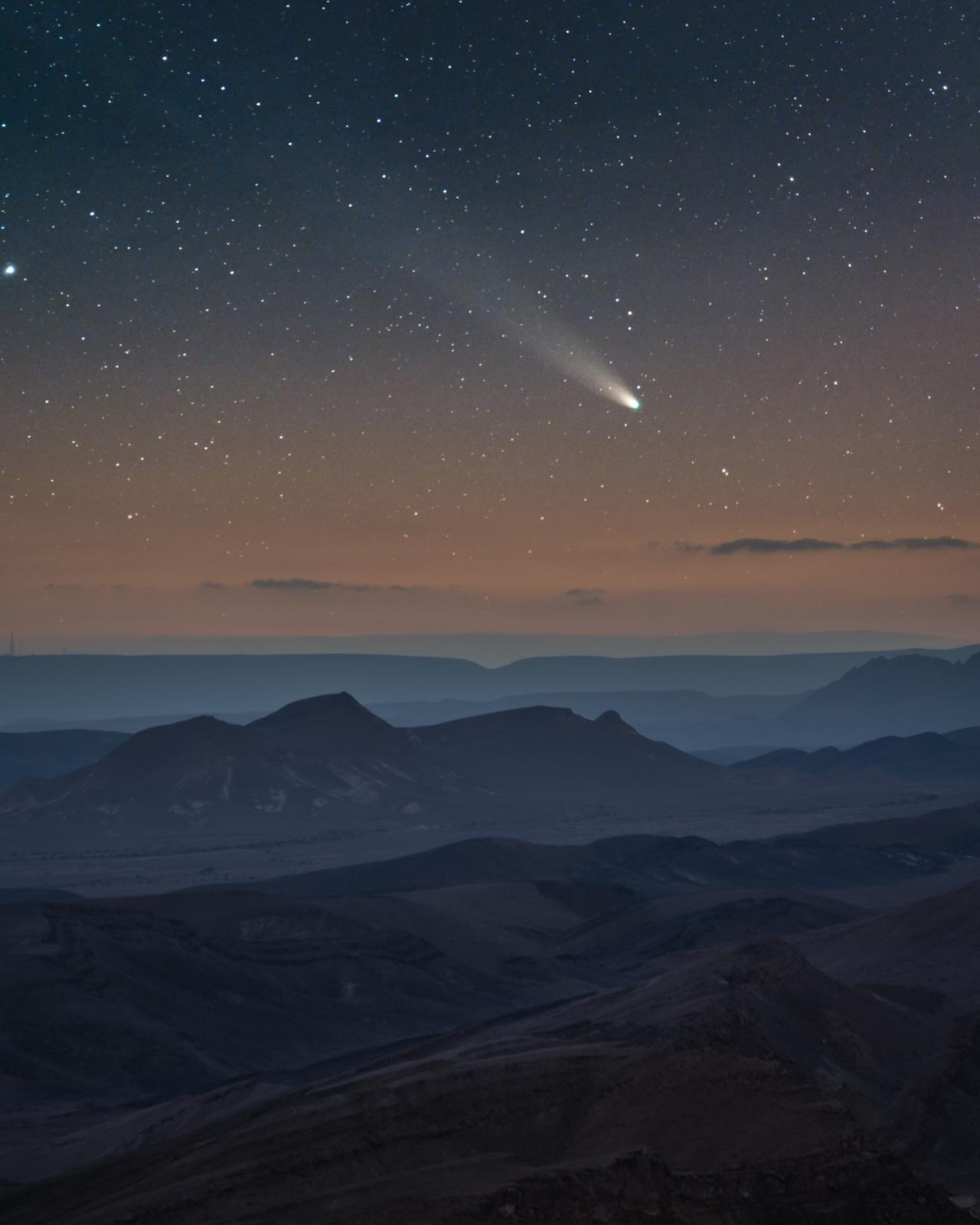Photograph of a comet entering the Earth's atmosphere. A rocky landscape occupies the bottom half of the frame, while the bright white tail of the comet can be seen moving across the sky above