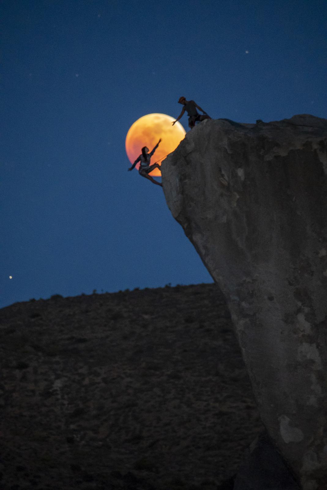 Silhouettes of rock climbers climb an overhanging rock with full Moon behind