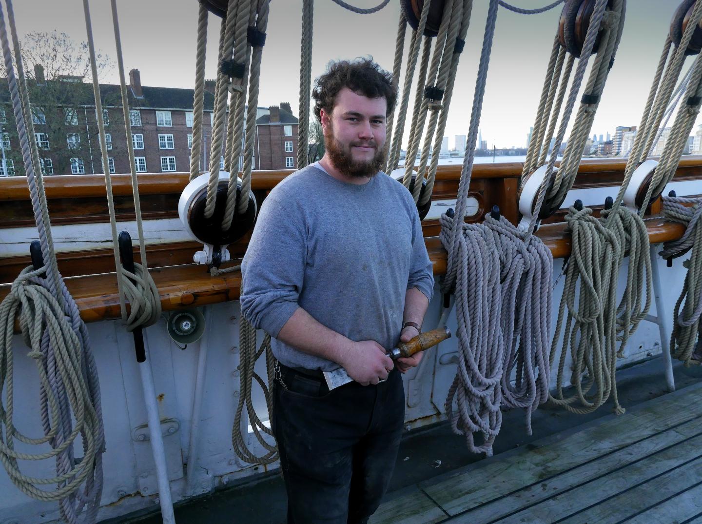 A man wearing a grey jumper stands on the deck of Cutty Sark holding a chisel