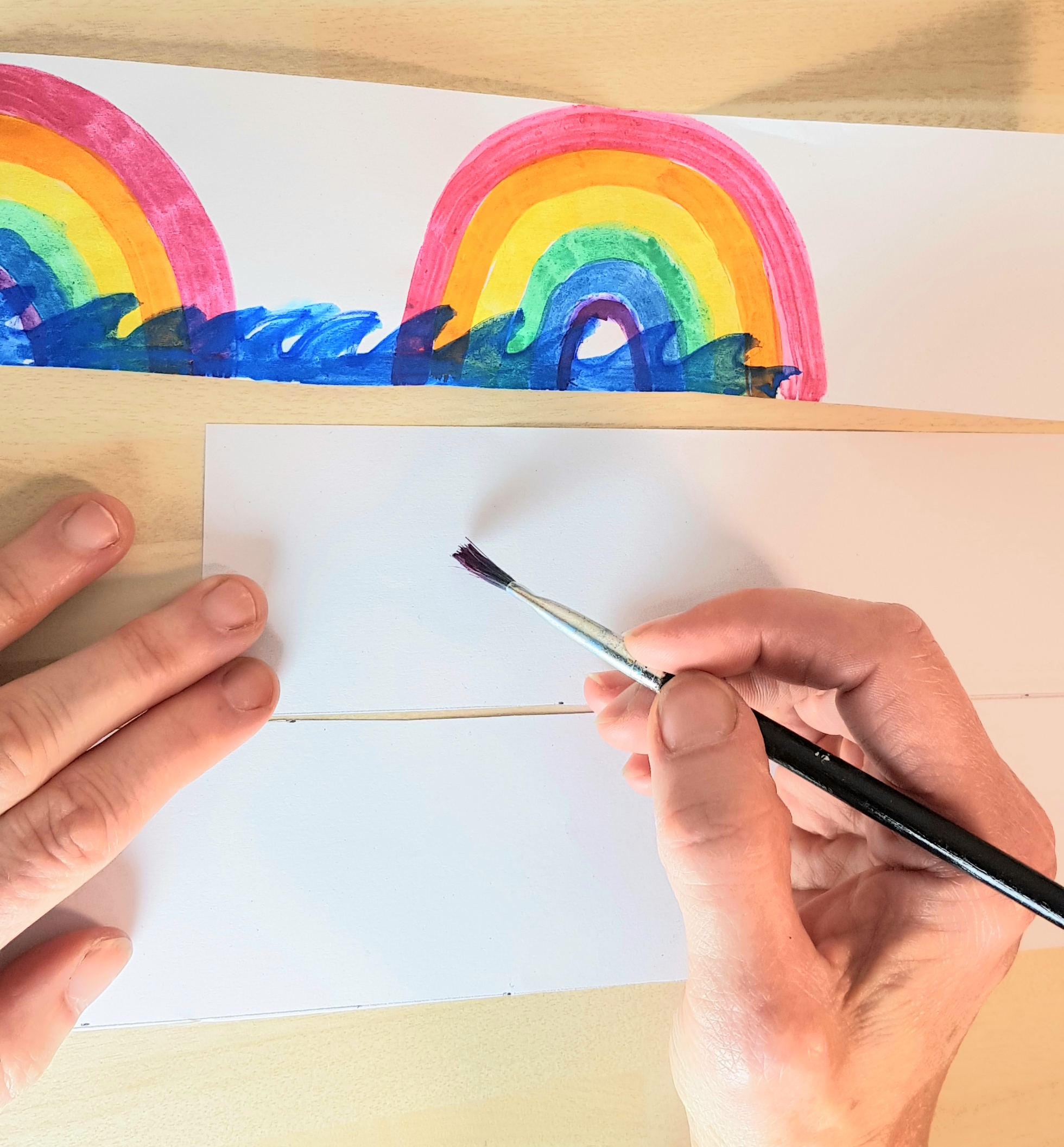 Decorate your strips of paper. 3 strips of paper have been cut out. One is decorated with rainbows and the ocean. Someone is preparing to start decorating the second strip of paper with a paintbrush.