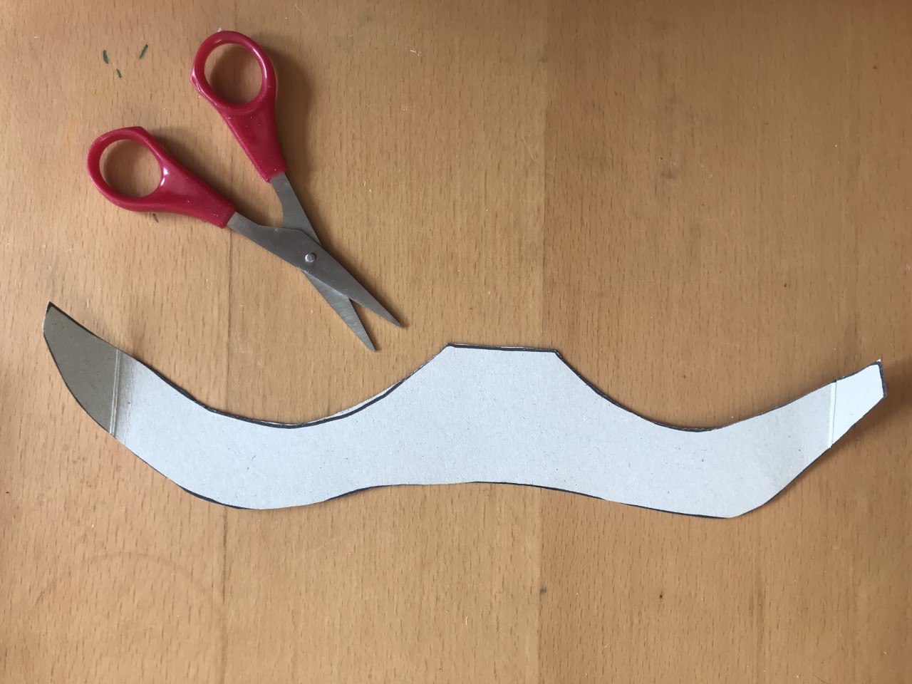 Cut out the wings A pair of red scissors are next to a piece of card cut out it a flattened W shape like a pair of wings.