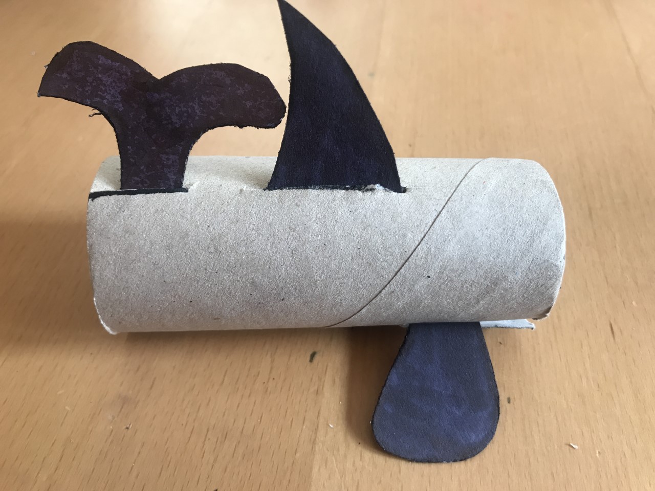 Make cuts and add your tail and fins A loo roll tube lies on it's side. A black whale tail and 2 fins have been added.