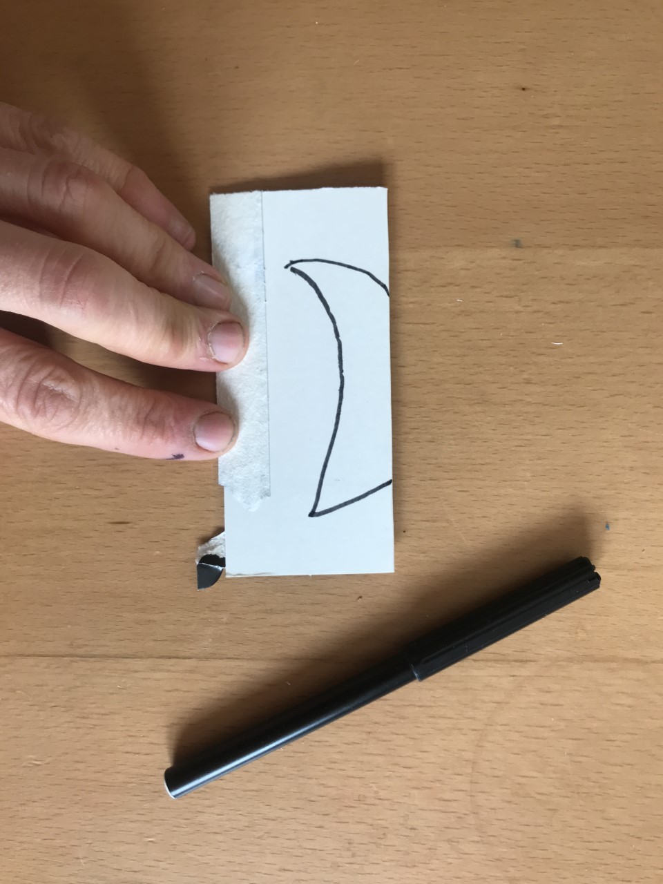 Draw this shape onto folded card Someone folds a piece of card. On it is drawn a crescent moon shape. The outer edge is against the edge of the cardboard. A black pen lies nearby.