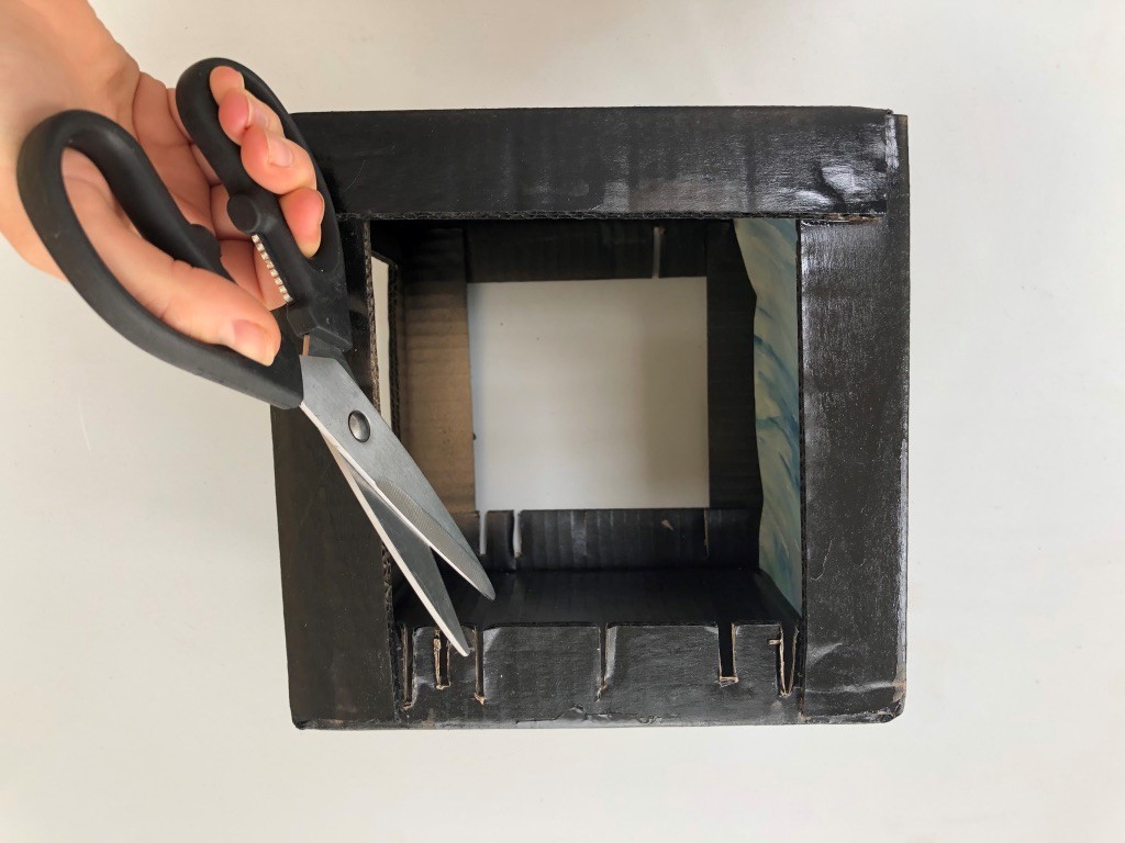 Make cuts in the bottom edges of your left and right windows. A cardboard box with windows cut on three side. Small cuts have been made in the two side windows.