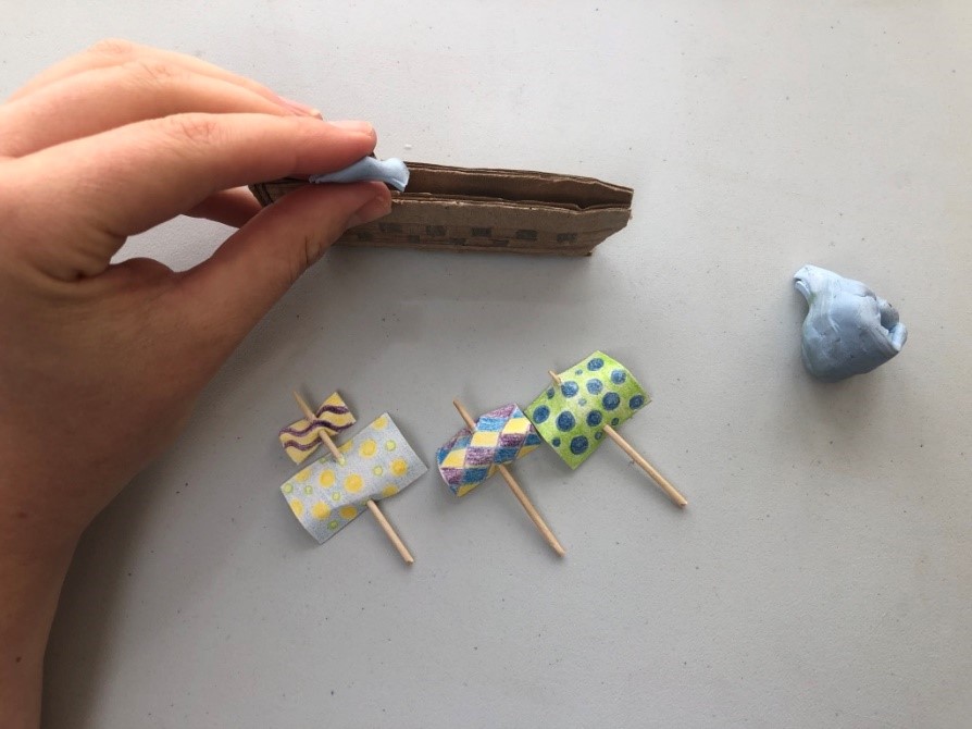 Add blue tac to your ship Someone puts a lump of blue tac inside a small ship made of cardboard. Three sails made of paper and toothpicks lie nearby.