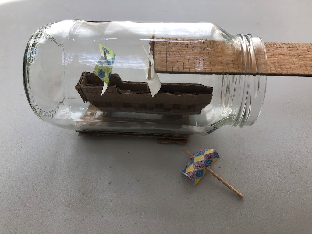 Put your sails in place Someone uses a ruler to place sails made of paper and toothpicks inside a ship. The ship is inside an empty jar