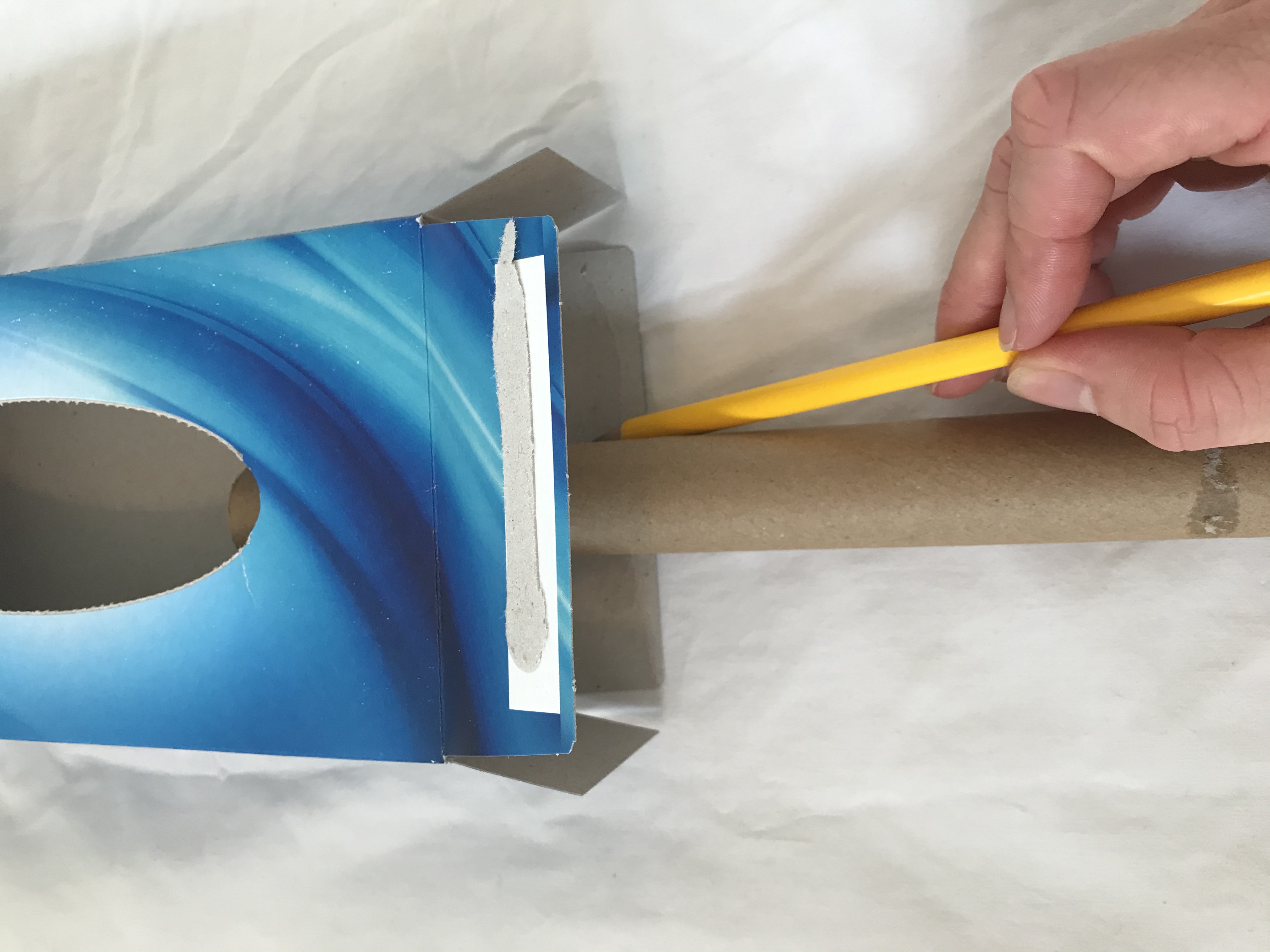 Mark where the tube is sat A cardboard tube is placed inside a tissue box. Someone is marking on the end of the box where the tube is.