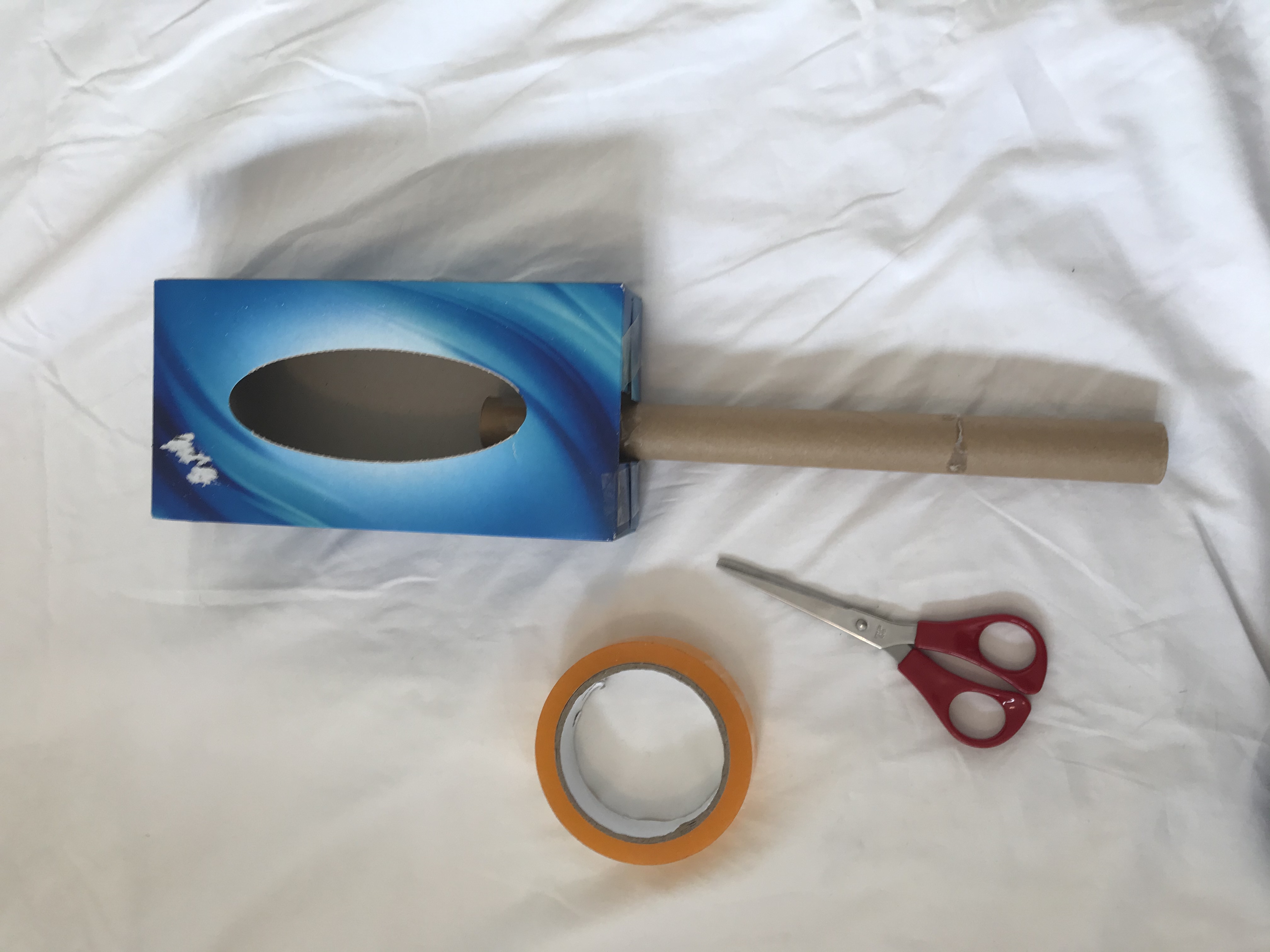 Seal up the end around the tube A tissue box with a cardboard tube attached to the end. A pair of red scissors and a role of tape are nearby.