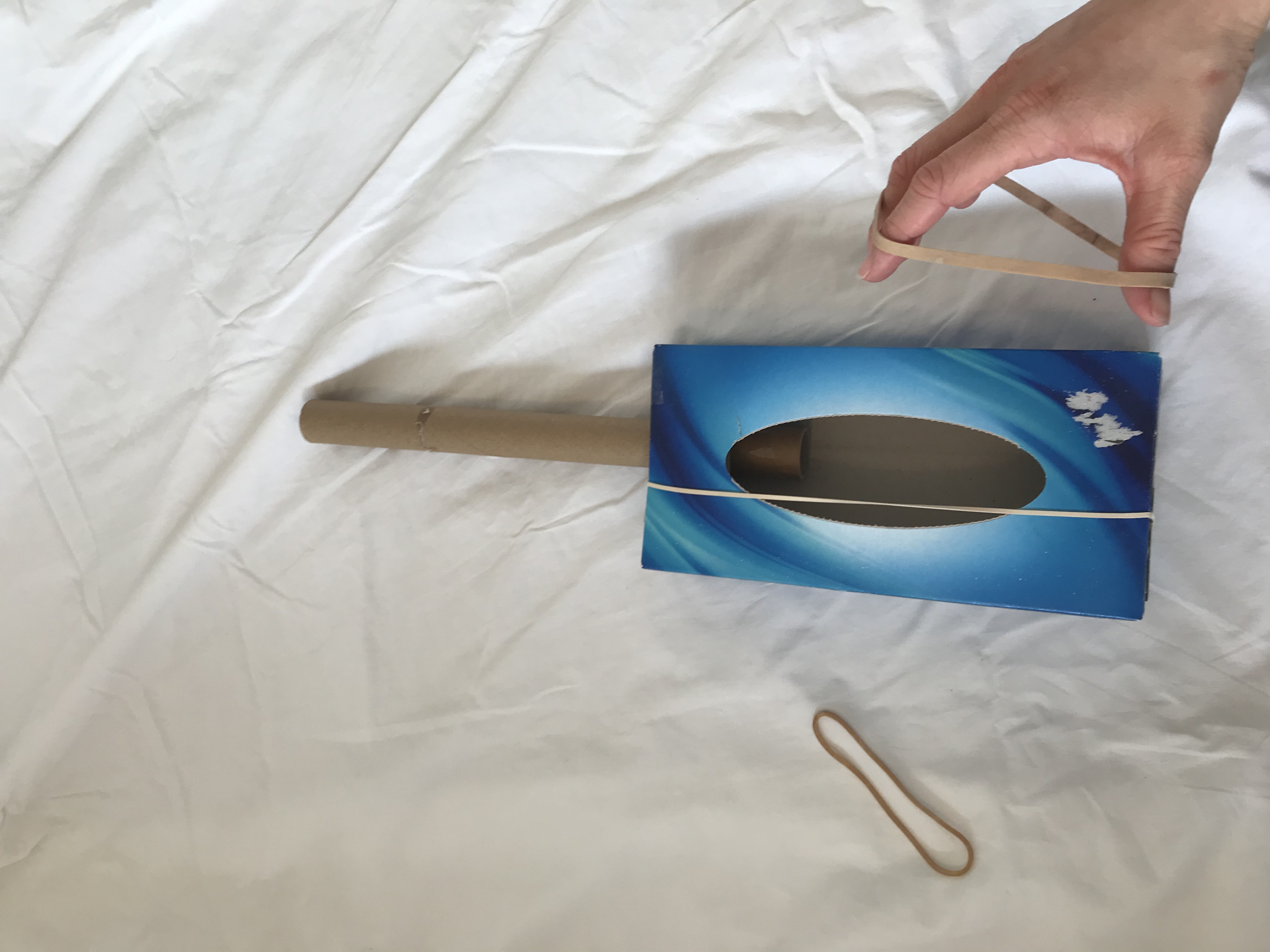 Add your elastic bands Someone adds elastic bands around the length of a tissue box. A cardboard tube has been stuck to the end of the box.
