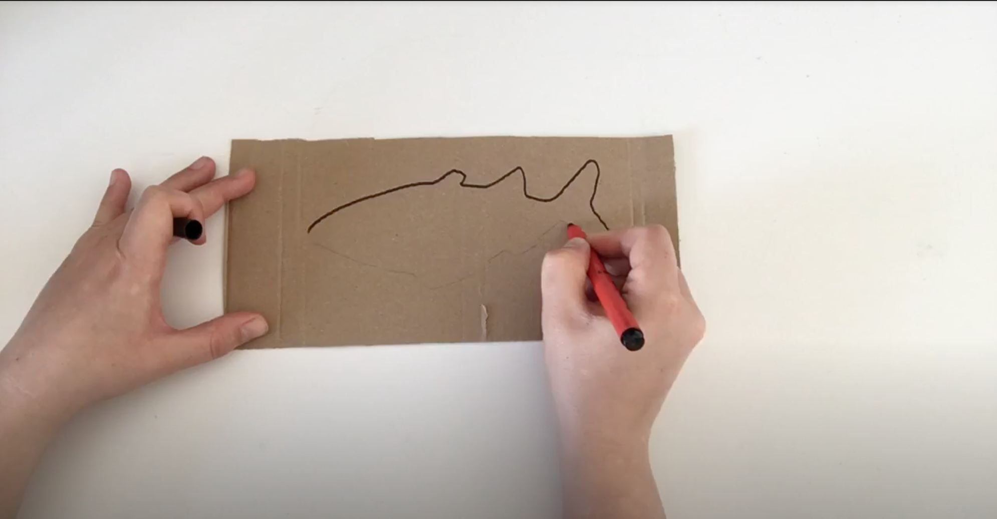 Image shows a hand drawing a fish onto a piece of cardboard Image shows a hand drawing a fish onto a piece of cardboard