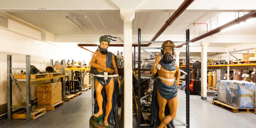 Inside Royal Museums Greenwich collections centre - rows of objects including two ships figureheads