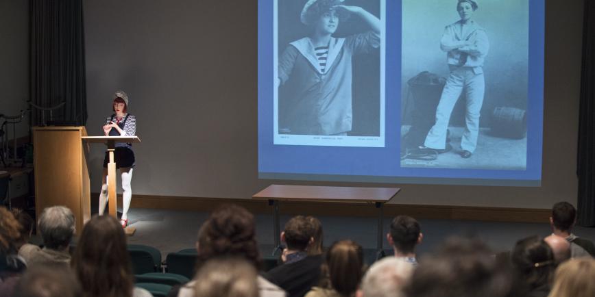 Photo of the Lecture Theatre at the National Maritime Museum filled with an audience watching a women present a talk on queer life at sea. Projected are two black and white photos of sailors, one wearing white bell bottoms and the other looking out to sea.
