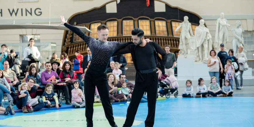 Photo of two male ballroom dancers  wearing black outfits, one with diamantes across his shoulders. A full audience sits behind the two dancers on the Great Map in the National Maritime Museum. 