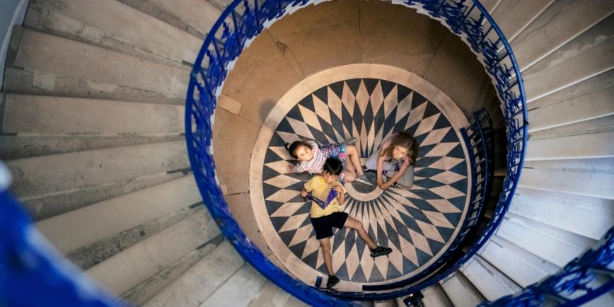 A group of children sit on the floor, looking up at a spiral staircase