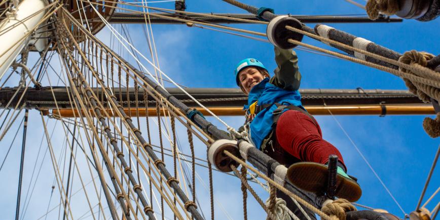 A woman climbs the rigging of historic sailing ship Cutty Sark. She is looking down at the photographer on deck, and the rigging and masts are above her