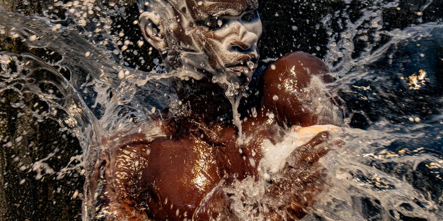 A photograph of a man washing. His face and hair is covered in soap suds, an water is being thrown over him