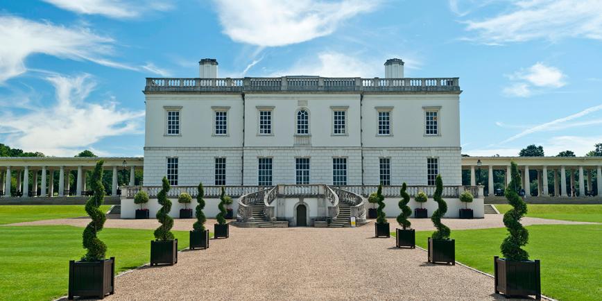 A view of the Queen's House in Greenwich from the outside. The square white building is almost symmetrical from the front, with a gravel pathway leading up to the main entrance