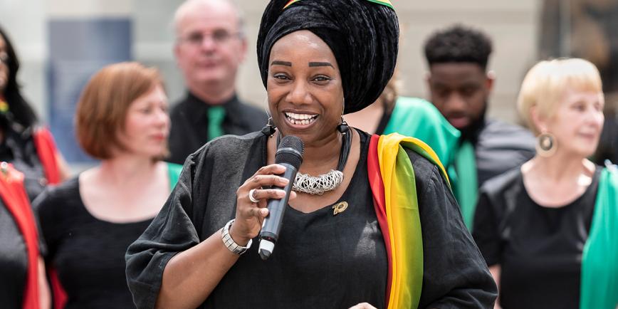 A female singer performs a solo as part of a choir performance. She is holding a microphone and is wearing a black dress with a multicoloured sash over her left arm, with a matching headscarf