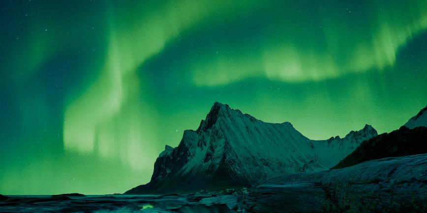 Image showing mountain and rocks in foreground, with rippling emerald aurora in the sky behind the mountain, which is reflected in a pool of water