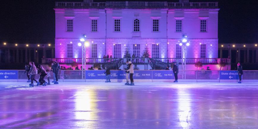 Image of the Queen's House Ice Rink with the classical building of the Queen's House in the background, lit up purple and blue, with a quite empty rink in front of it, only around 8 people on it