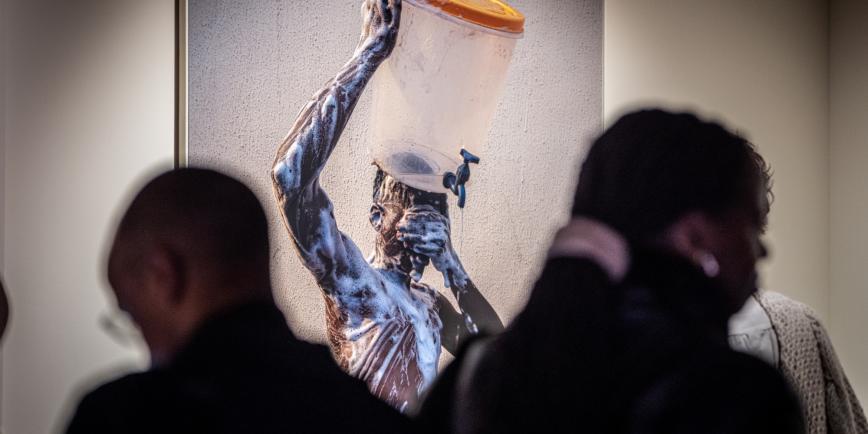 Guests in shadow walk around a contemporary art exhibition. On the wall in the background, lit up brightly, is a striking photograph of a young black man washing. He is balancing a bucket on his head with one hand and covering his eyes with the other. His body is covered in soap