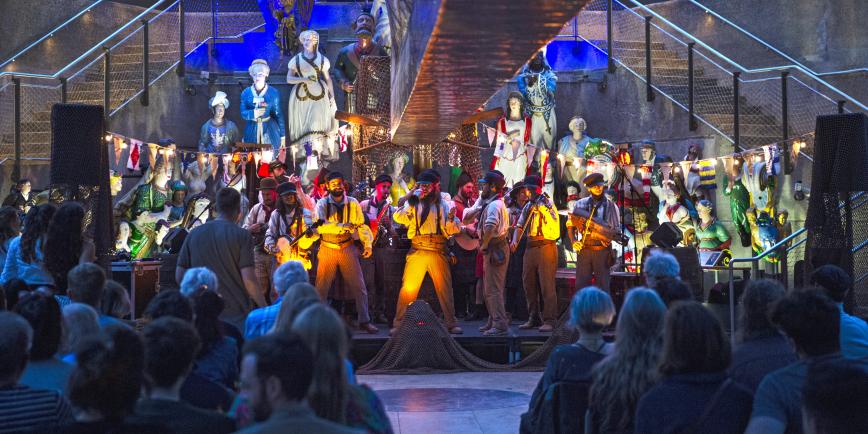 Image of band of people under the copper hull of Cutty Sark sailing ship, standing on a stage wearing shirts and paper bag trousers, holding instruments. Behind them are an array of ship's figureheads.