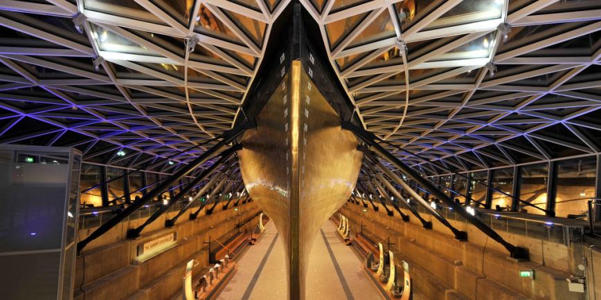 Photo of a large copper hull of a ship in a 'Dry Dock', a dock with stepped sides and supports holding up the ship. The roof is made up of loads of triangular glass panels.