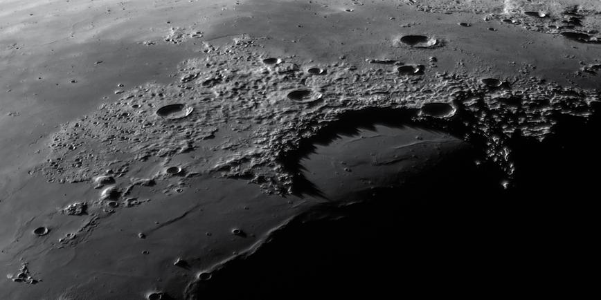 Image showing a close up of the surface of the moon from a diagonal angle looking down from just above it, with the curve of the moon visible in the top of the image. The lunar surface is pockmarked with many round craters of different sizes and depths, and raised areas showing hills and mountains on the surface. The image fades to blackness in the bottom right hand corner.