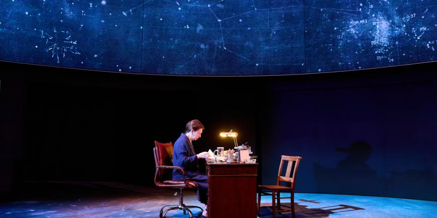 A scene from theatre production The Lightest Element. A woman sits alone at a wooden desk in the middle of the stage, with dramatic lighting evoking a night sky filled with stars