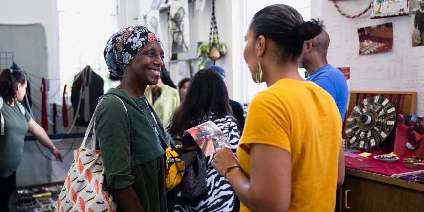 Two women converse happily during an activity at a Museum. One is wearing a patterned headscarf and has a tote bag over her shoulder; the other has a yellow top and is facing away from the camera