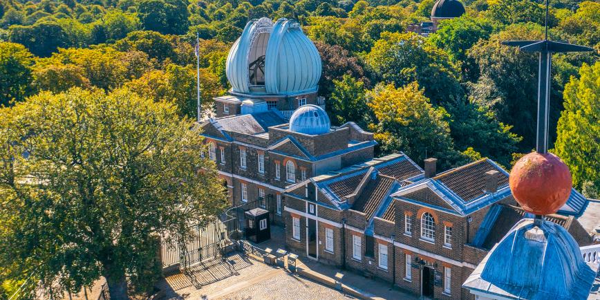 Drone view of the historic Royal Observatory buildings during the day. Trees surround a central group of brick buildings, with the distinctive 'Onion Dome' of the Great Equatorial Telescope in the centre
