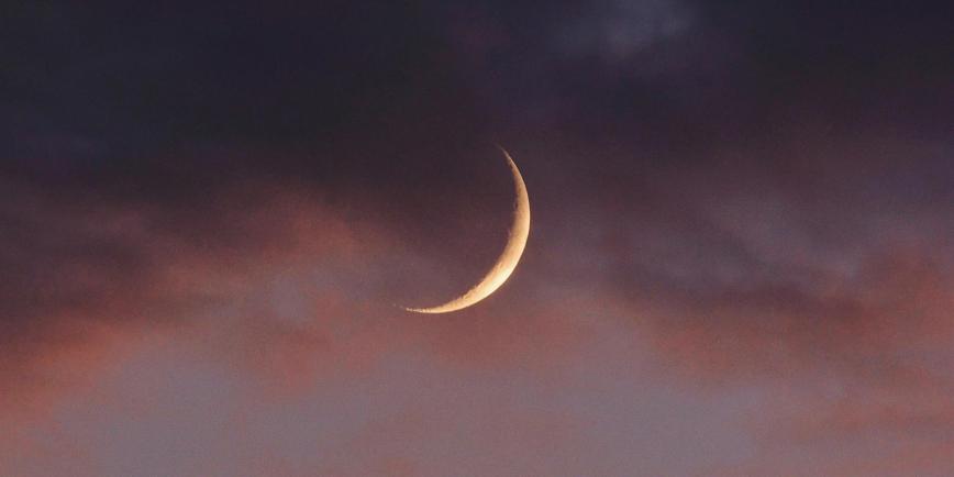 The new crescent Moon in the evening sky, surrounded by pinkish clouds and a darkening sky