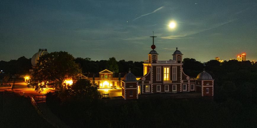 A drone photograph of the Royal Observatory at night, the Moon is bright in the sky and lights in the observatory buildings below casts a yellow glow