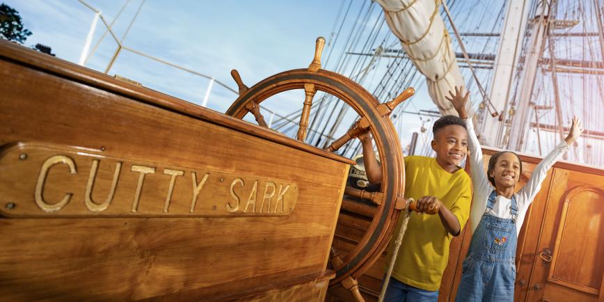 Photo of two children on board a historic sailing ship next to the ship's wheel looking happy and excited
