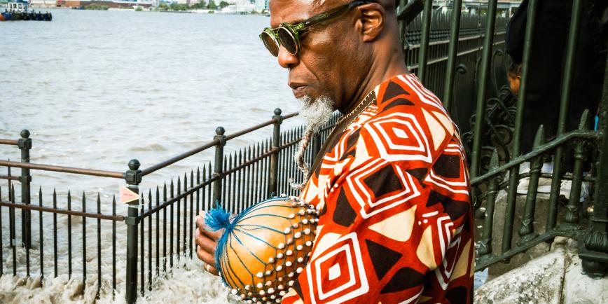 A Black man wearing green sunglasses and a geometric patterned top stands contemplatively by the River Thames