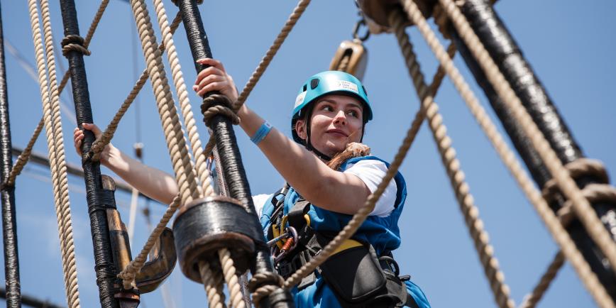 A woman gazes confidently out as she holds on to the rigging of a tall sailing ship
