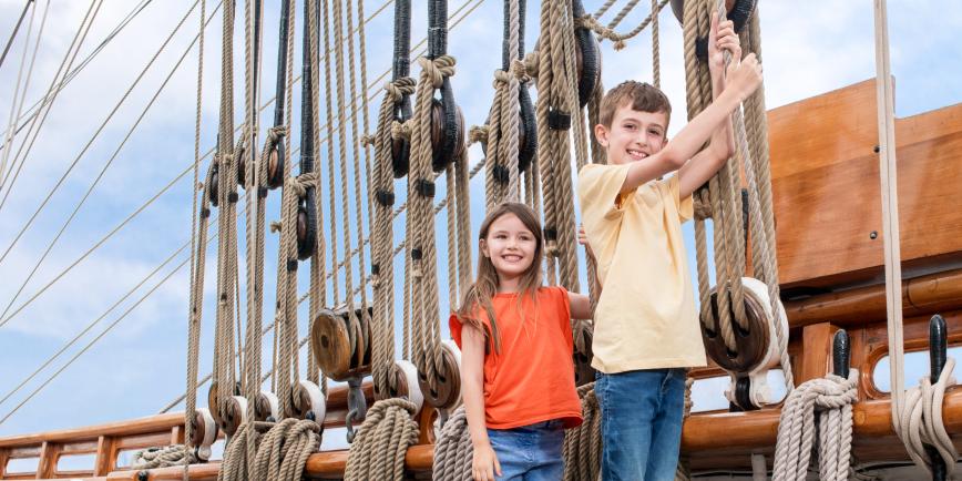 Photo of kids on a historic ship, a younger girl and older boy holding onto ropes that make up the rigging of the ship and smiling
